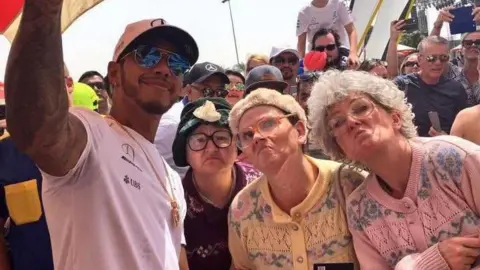 Fizzog Productions Three women wearing knitted jumpers and grey curly wigs are standing next to Lewis Hamilton, who is wearing a white t-shirt with a Mercedes logo and a white cap on his head. Crowds surround them as they take a selfie together. 