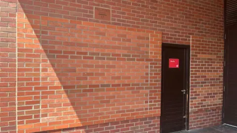 A wall of brown bricks. More than 100 of the bricks have names written on them. A larger block at the top of the section has Wall of Fame Panel S5 written on it.  There is a black door next to the panel, with a red media sign on it. 