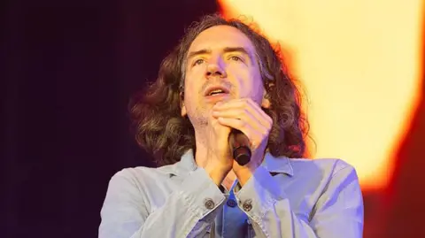 Getty Images Gary Lightbody - a man with dark hair past his shoulders - holds a microphone while looking out from the stage at a music festival