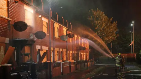 PA Media Image of two fire fighters spraying a terraced house with water, the windows are boarded up and it is dark outside, with a red glow from nearby flames