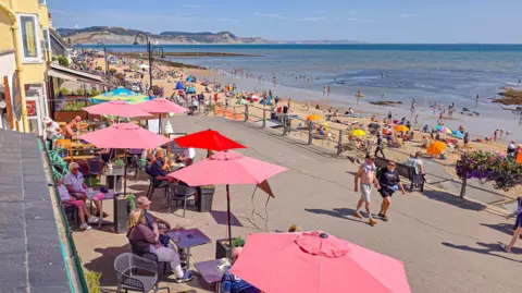Love Lyme Regis View looking down on cafe tables and red umbrellas outside a cafe on the promenade at Lyme Regis. It's a sunny summer's day and people are sitting at the tables, walking along the promenade and the beach and shore is packed with people.