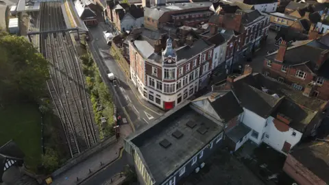 An aerial drone shot of a large red brick corner building with white frontage and windows. It has a clock tower and a spire. To its left are railway tracks