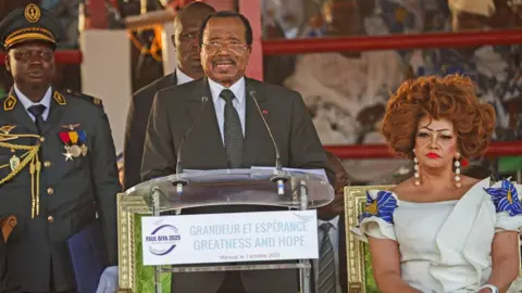 Reuters President Paul Biya speaks at a lectern dressed in a suit. His wife Chantal sits to his side in a white dress with floral ornaments on the shoulders. Behind them stands a military officer, another official and behind them supporters of Cameroon's president - Maroua, Cameroon, 7 October 2025.
