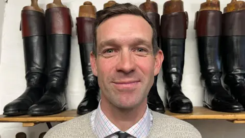 Daniel is looking directly into the camera and smiling slightly - he's wearing a beige jumper with a check shirt and black tie underneath. Behind him is a rack of black riding boots, like those worn for hunting. 