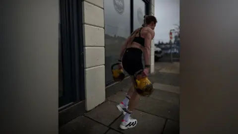 POW8R A woman dressed in black shorts and workout top and white trainers is carrying two kettlebells in the street outside a gym in Liverpool