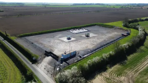A large concreted area, rectangular in shape, is surrounded by security fencing. It sits in the middle of flat farmland.