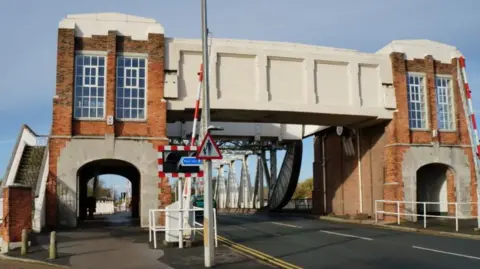 Ian S/Geograph A view of the bridge from the pavement. It has a huge metal structure and brick towers either side with archways for pedestrians to walk through.