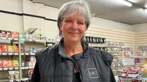 BBC/JULIA LEWIS A woman with short grey hair and wearing a black top and black gilet, with an L&C logo on the chest, is standing in front of rows of groceries.