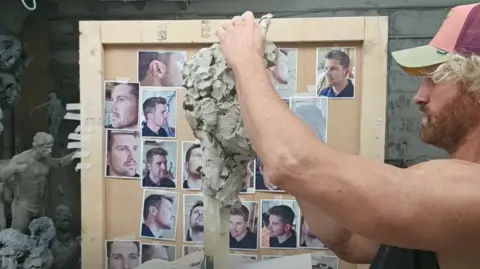 BBC/Chatterbox Media Nick Elphick has blond curly hair coming out of the sides of his baseball hat. He is looking at a clay model base and moulding it with his hands, with a pinboard of photos of Frost in the background.
