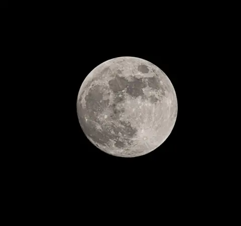 Andrew McIntosh The full Moon appears centred in the frame, with its surface features—craters and grey lunar maria—clearly visible against a stark black background.