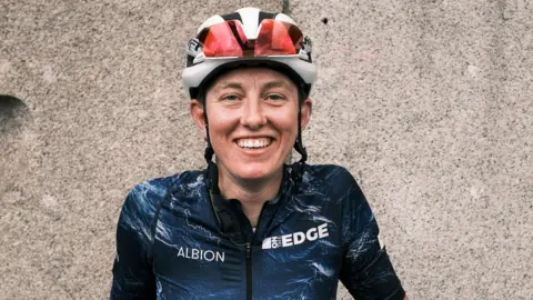 A woman wearing a red and white cycling helmet and a sponsored dark blue cycling top, smiles while standing in front of a stone wall.