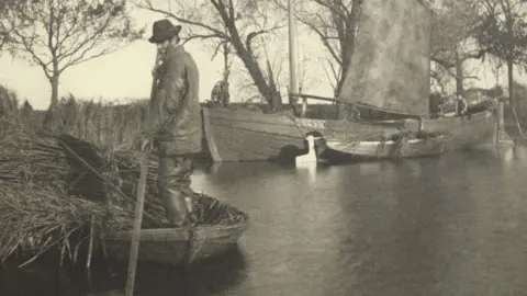 Black and white image of a man pushing through a river on a small boat loaded with reeds with a small wherry boat in the background