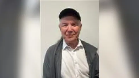 City of Doncaster Council Image of Mark Broadhurst from the waist up. He is wearing a white shirt and a grey jacket as well as a black cap. He is smiling at the camera.