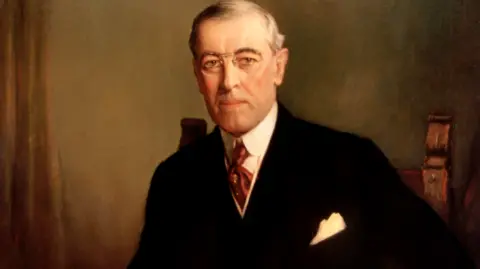 Frank Graham Cootes Image shows the official portrait of Woodrow Wilson. He is wearing a black suit, white shirt, burgundy tie and wire-rimmed glasses. 