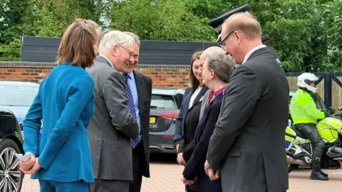 The Duke of Gloucester with white hair wearing in a grey jacket is second from the left next to a woman in a blue jacket on the left. Five people are on the right and cars are in the background.