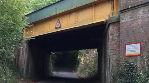 Broom Lane Bridge pictured in high summer with the trees in full foliage, the lower girder painted yellow and the upper girder painted green.