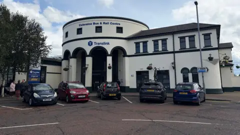 Coleraine train station, a large white building with cars parked outside. A sign on the top of the building reads "Coleraine Bus & Rail Centre" in navy blue font. Further down there is a Translink logo in the same colour.