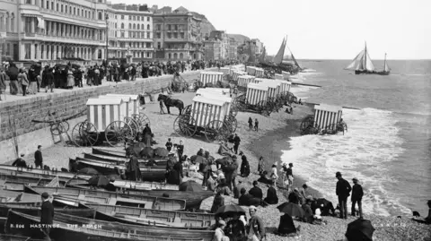 A black and white photo of horses and carriages on Hastings beach as well as people.
