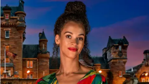 Roxy is in series four of The Traitors, wearing a green and red shirt and red lipstick with her hair piled high on her head, standing in front of the Traitors castle