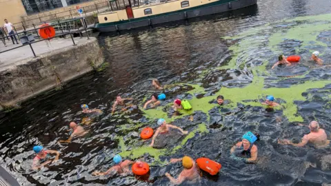 Steve Jones/BBC A groupd of about a dozen people swimming in an area of open water at Leeds Dock. A narrowboat can be seen in the background.