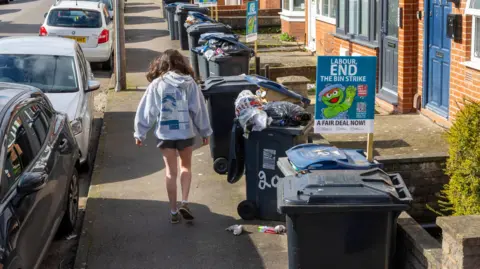 A woman walks down a Birmingham street seen with overflowing wheelie bins on one side of the pavement and parked cars on the other