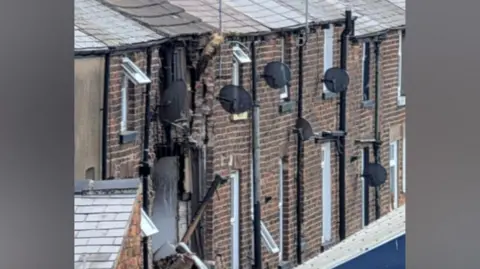 The image shows the rear side of a row of older brick terraced houses, viewed from an elevated or slightly angled perspective. The focus is on a section of severe structural damage affecting one of the properties.
In the centre of the image, one house has extensive vertical damage running from the roofline downward.
A large section of the brick wall has collapsed or been blown outward, exposing the interior structure.