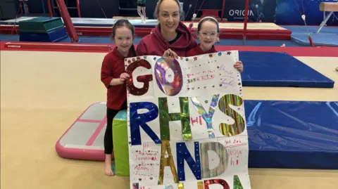 Woman in gym holding a poster in support of Rhys McClenaghan, wearing red top, flanked by two girls in red.