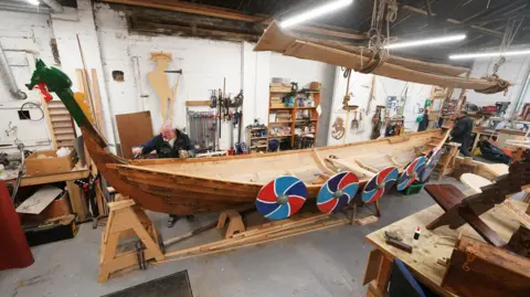 Two men are working on a Viking boat in a boatshed. It is about five metres long, narrow with a figurehead and blue and red circles attached to the side.