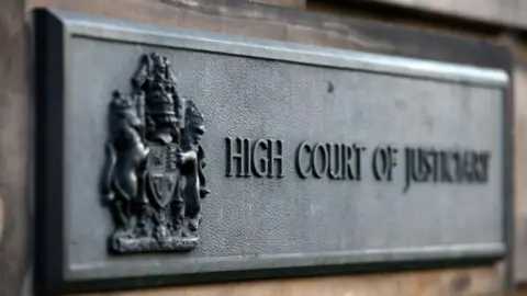 A sign attached to a wall which says High Court of Justiciary with a crest