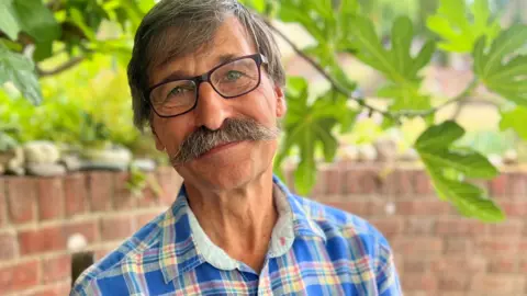 Dr Alain Gregoire wearing a blue checked shirt sitting in front of a low brick wall with overhanging trees behind