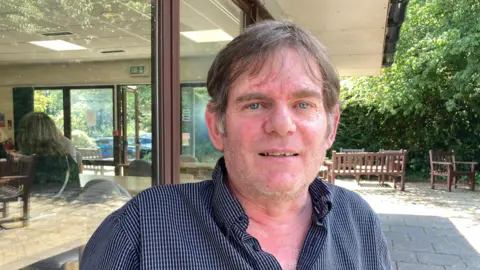 BBC Close up of Kevin Heard's face, close to a building's windows. He has brown hair, blue eyes and is wearing a blue shirt with the collar unbuttoned.