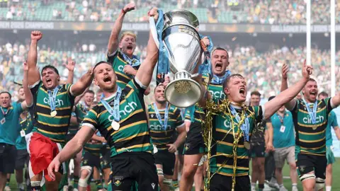 Northampton Saints players cheer as they carry a huge trophy, they are wearing medals and gold confetti