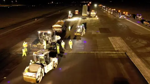 BBC/STEVE HUBBARD A still taken from a drone looking down at the runway at London Luton Airport. It is night and the runway is well lit with a series of construction vehicles in the foreground, including a roller and tipper trucks - one of which is tipping out material. The scene includes eight workers dressed in yellow high-visibility outfits. To the right of the picture is a long line of vehicles. 