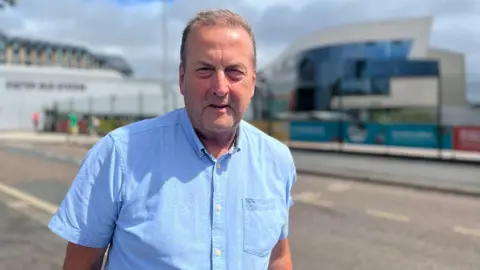 A man wearing a short sleeved blue shirt is standing on a pavement outdoors. Buildings are visible in the blurred background behind him. 