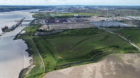An aerial shot of the Port of Tilbury, which has lots of containers stored on the banks of the River Thames. The shot shows a large green field next to a muddy brownfield site, both of which will be used to build Tilbury3.