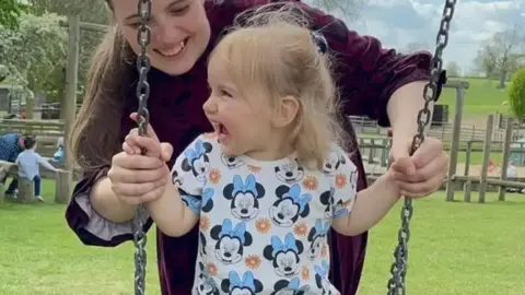 Instagram Isabella smiling on a swing with her mother smiling and standing behind her. Isabella has a T-shirt on with Minnie Mouse's face on it.