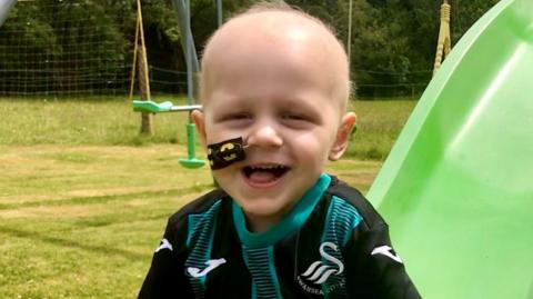 Joseph Yeandle smiling while playing on a slide in a park. He is wearing a replica Swansea City shirt, with a tube related to his treatment attached to his face going into a nostril