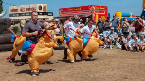 Nadine Hudson-Featherstone Men wearing rodeo horse costumes dancing in front of a crowd of people.