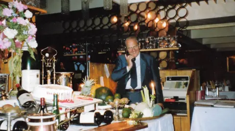 Family Photo Eddie Rabaiotti stands smiling with a phone held up to his ear. Behind him is a wooden shelf holding the till and in front of him is a table laden with wine bottles, candles, a cake and an assortment of fruit including limes, pineapples and kiwis. There is also a bouquet of flowers on display. 