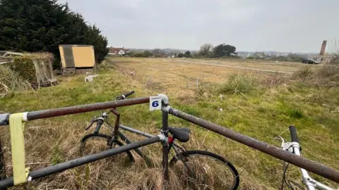 Some rusty bikes chained up by a scruffy field.