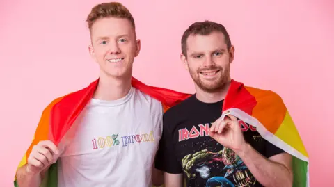 Comedians James Barr and Dan Hudson pose with a Pride flag to promote their podcast "A Gay and a NonGay".