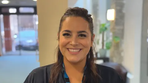 George King/BBC A woman with long brown hair tied back smiling into the camera