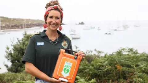 Grace stood holding the orange defibrillator. She is stood in front of a sea view which has boats on the water. 