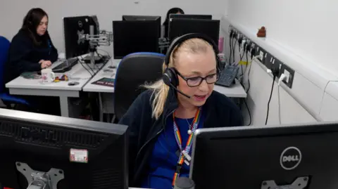 Three women wearing telephone headsets sit behind computer monitors in an office with white walls. Closest to the camera, a woman with blond hair, tied back, talks into her headset as she looks at her screen. She is wearing dark-rimmed glasses, a navy-blue fleece, a mid-blue top and a rainbow-coloured lanyard with "NHS" logos on it. Behind her a woman with long dark hair, wearing a black fleece, is typing at a keyboard. Another woman is largely hidden by a monitor.