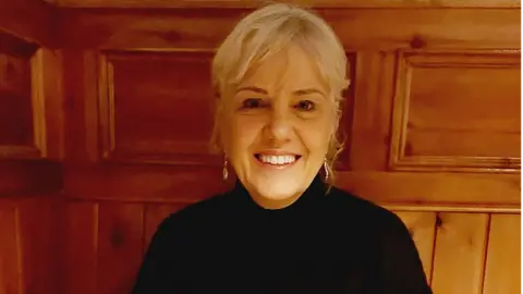 Jenny Dunlop. She has blonde hair tied back with a fringe, wearing earring and a black top. Wood panelling is behind her.