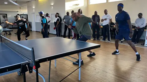 Nicola Haseler/BBC A group of men playing table tennis in a room with a wooden floor. About five men are playing and the rest are standing behind. The table tennis tables are blue. There are small high up windows behind them. 