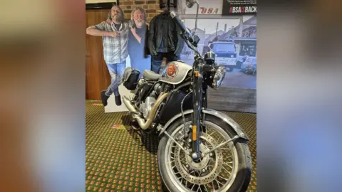 The BSA Gold Star pictured at The National Motorcycle Museum. It has a silvery-grey, chrome and black colour scheme. Behind it is a carboard cut-out of the Hairy Bikers and a leather jacket.