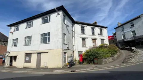 A white building is positioned at a road corner, with a red mail box in front of it. There is a blue sky seen above and behind it. 