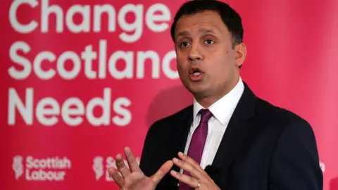 PA Media Anas Sarwar, wearing a dark suit and purple tie, speaks in front of a red Scottish Labour sign in a medium close-up shot 