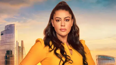 Picture of Nadia Suliaman in a mustard yellow top. She stands infront of a green screen of London. She looks blankly into the camera and has brown eyes and dark brown hair. 